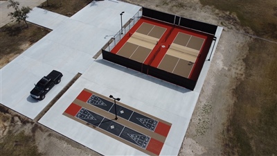 Outdoor sports area with two sections: one with a beige and red surface housing two tennis courts and one with multiple shuffle boards for multipurpose uses. 