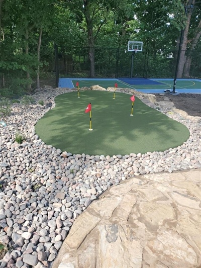 Outdoor residential area featuring a small putting green with two red flags, surrounded by rocks and trees, with a blue sports court and basketball hoop in the background in Wayzata.