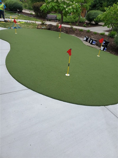 Outdoor putting green with several red flags, surrounded by a concrete pathway and lush greenery.