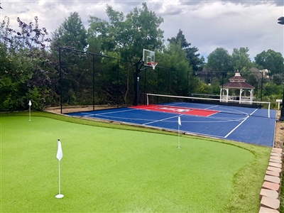 Outdoor residential area featuring a green putting green with white flags, a blue and red sports court with a basketball hoop and tennis net.