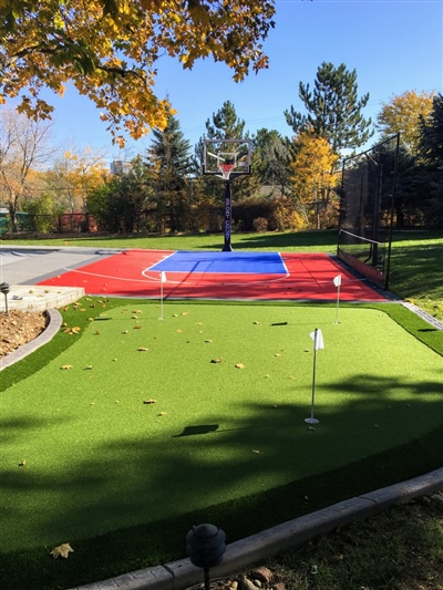 Outdoor residential area featuring a green putting green with white flags, and a red and blue sports court with a basketball hoop.