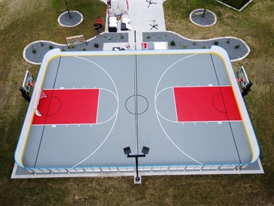 Outdoor multi-sport court featuring two red basketball areas within a grey hockey rink, surrounded by a fence.