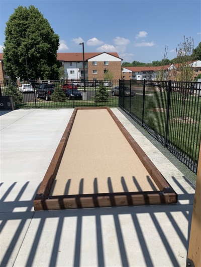 Outdoor bocce ball court with wooden borders, located next to a concrete walkway and fenced area.