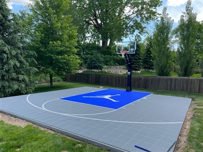 A backyard basketball court in Wayzata, MN featuring durable outdoor basketball flooring, surrounded by lush green trees and a wooden fence, with a blue Jumpman logo at center court in Oconomowoc.