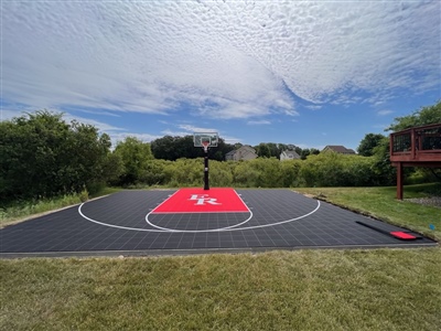 A backyard basketball court featuring premium outdoor basketball flooring in black and red, with a FR logo in the paint, surrounded by lush greenery and clear blue skies.