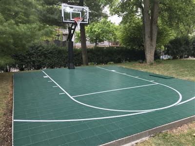 A basketball court with a green surface, white lines, and a basketball hoop in a wooded backyard in Richfield.