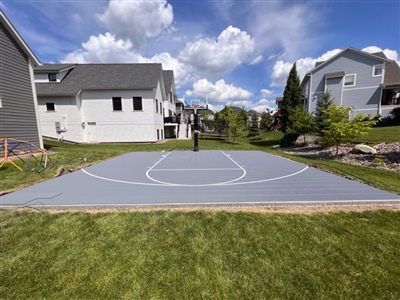 A basketball court with a grey surface and white markings with a basketball hoop in a residential area.