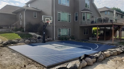 A residential basketball court with a blue and grey surface and white lines surrounded by a rock retaining wall.