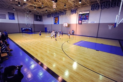 A basketball court with a maple wood-like surface, black lines, and a sport court logo in the center with kids playing basketball..