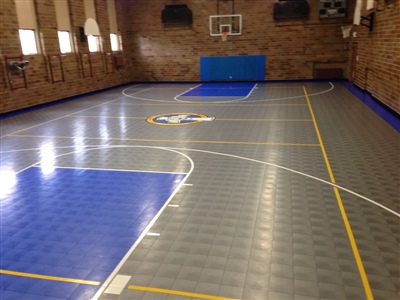 An indoor basketball court with a grey and blue surface, yellow lines, and a central logo, set against brick walls.