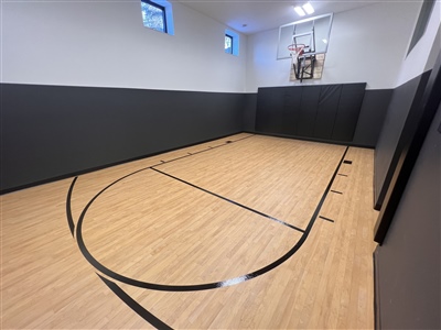 A small indoor basketball court with a polished wood surface, black lines, and grey walls, featuring a basketball hoop at one end.