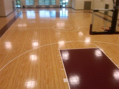 An indoor basketball court with a polished wood surface, maroon key areas, and white lines, featuring a central logo.