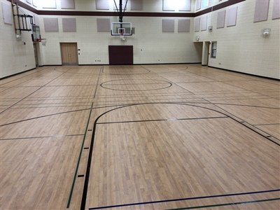 An indoor basketball court with a light wood surface, black lines, and white walls, featuring a basketball hoop at one end and bright overhead lighting.