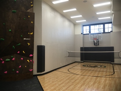  A small indoor basketball court with a light wood surface, black lines, and white walls, featuring a basketball hoop and a rock climbing wall on one side in Plymouth.