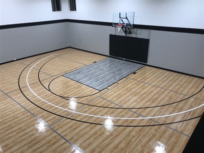 A small indoor basketball court with a polished wood surface, black lines, and grey walls, featuring a basketball hoop and a protective mat in the center.