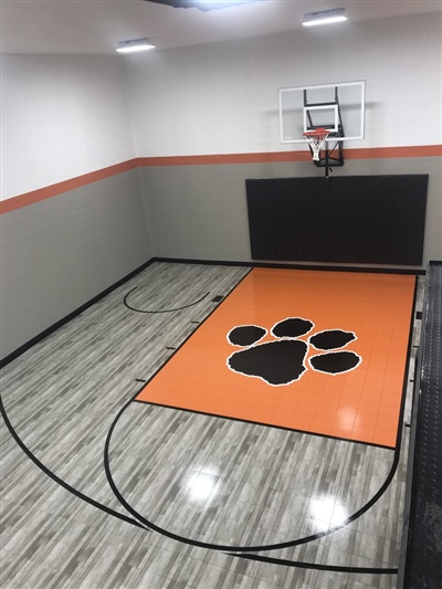 A small indoor basketball court with a grey wood-like surface, black lines, and light grey walls, featuring an orange key area with a black paw print emblem and a basketball hoop with black padding behind it in Farmington.