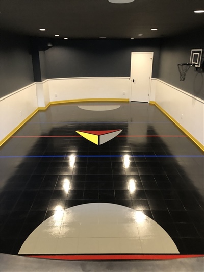 A small indoor basketball court with a black surface, white and blue lines, and dark grey walls, featuring a central emblem with red, yellow, and white colors.