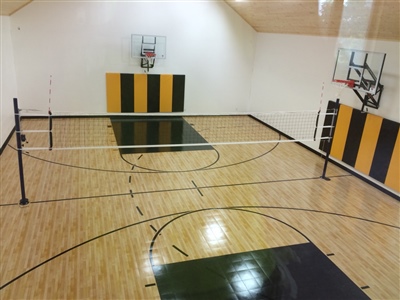  A small indoor basketball court with a polished wood surface, black lines, and white walls, featuring green and yellow padded areas behind the basketball hoops and a volleyball net set up across the court.