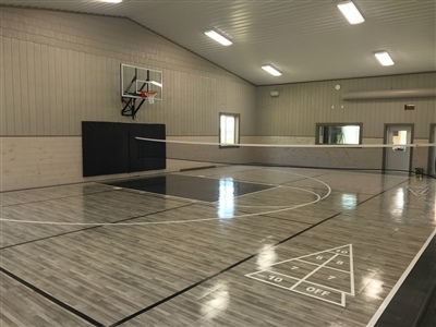 A small indoor basketball court with a grey wood-like surface, black lines, and light grey walls, featuring a black key area, shuffleboard markings, and a basketball hoop with black padding behind it.