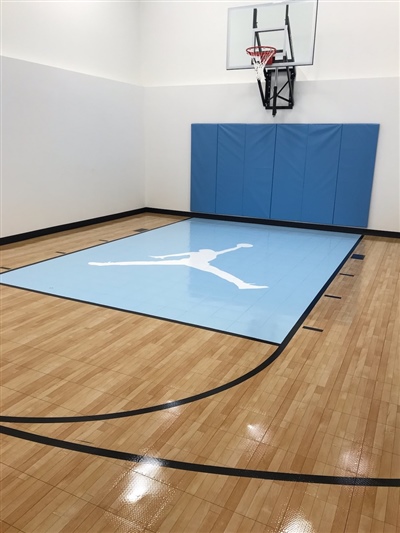  A small indoor basketball court with a polished wood surface, black lines, and white walls, featuring a light blue key area with a white Jumpman logo and a basketball hoop with blue padding behind it in Roseville.