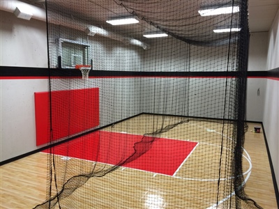 Indoor home basketball court with light wood flooring, red and black wall padding, a basketball hoop, and a black net hanging from the ceiling in Inver Grove Heights.