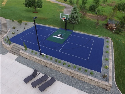  Outdoor residential sports court with a blue and green surface, a basketball hoop, surrounded by a stone retaining wall, and greenery in the background in Cedar Lake.