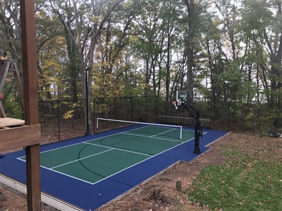 Outdoor residential sports court with a blue and green surface, a basketball hoop, and a tennis net, surrounded by trees and foliage.