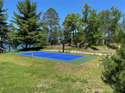 Outdoor residential sports court with a blue and green surface, a basketball hoop, and a tennis net, surrounded by trees and near a lake in Prior Lake.