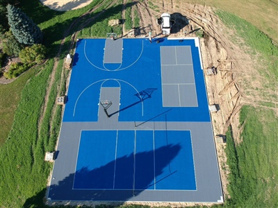 Outdoor residential sports complex with blue and gray surfaces, featuring two basketball courts and two tennis courts, surrounded by grass and trees in Woodbury.