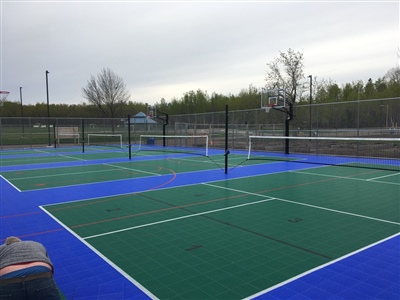  Outdoor sports court with a blue and green surface, multiple tennis nets, and surrounded by a fence and trees.