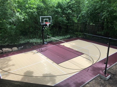 Outdoor residential basketball court with a beige and maroon surface, a basketball hoop, and surrounded by dense trees and greenery in Ham Lake.