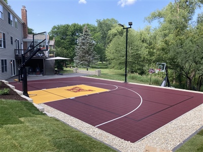 Outdoor residential basketball court with a maroon and yellow surface, a U of M logo in the center, and two basketball hoops.