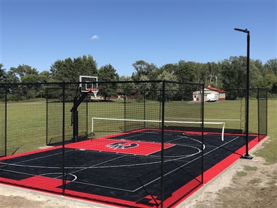 Outdoor residential sports court with a black and red surface with a logo in the center, featuring a basketball hoop and a tennis net, surrounded by a black fence and open grassy area.