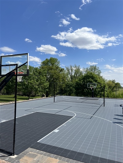 Outdoor residential sports court with a light and dark grey surface, featuring a basketball hoop and surrounded by trees and greenery in Andover.