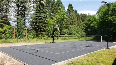 Outdoor residential sports court with a grey surface and white and black lines, featuring a basketball hoop and a tennis net, surrounded by trees and greenery.