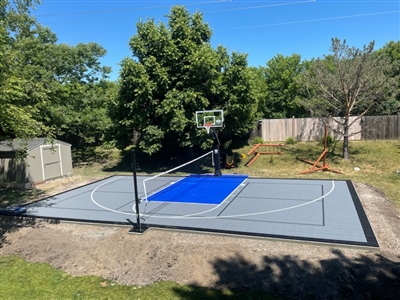 Outdoor residential sports court with a gray surface and a blue painted key area, featuring a basketball hoop and tennis net, and surrounded by trees and a fence.