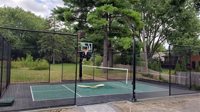 Outdoor residential sports court with a black and green surface, featuring a basketball hoop and a tennis net, surrounded by a black fence, trees, and greenery in Coon Rapids.