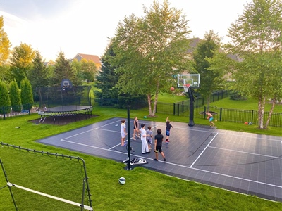 Outdoor residential sports court with a black surface, surrounded by greenery and a trampoline with kids playing basketball.