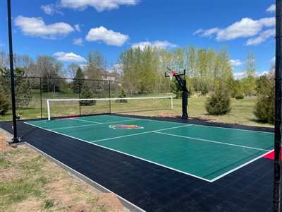  Outdoor residential sports court with a green and black surface, featuring a basketball hoop and a volleyball net, surrounded by trees and greenery.